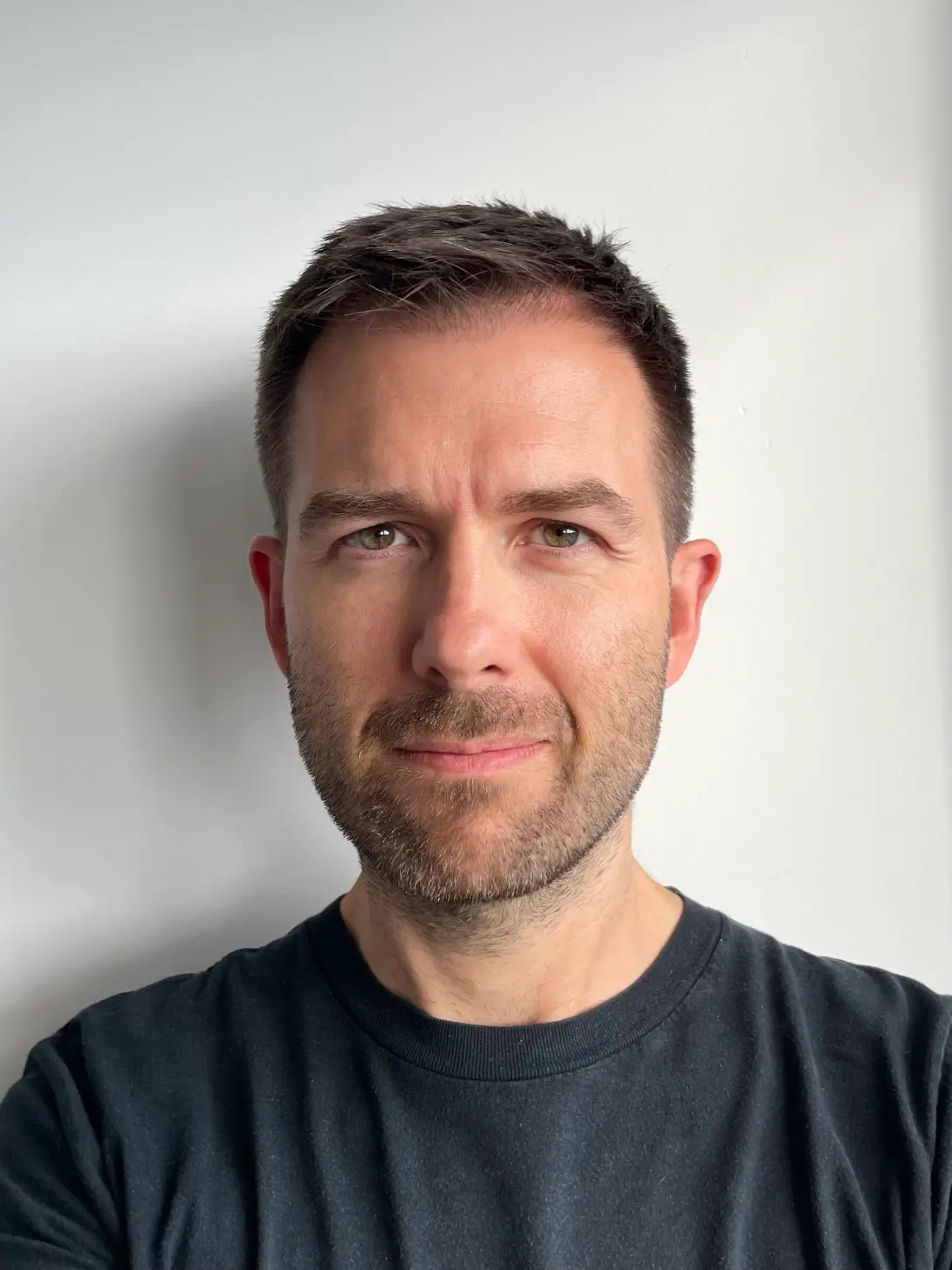 Man with short brown hair, light beard, and a black t-shirt standing against a plain white wall.