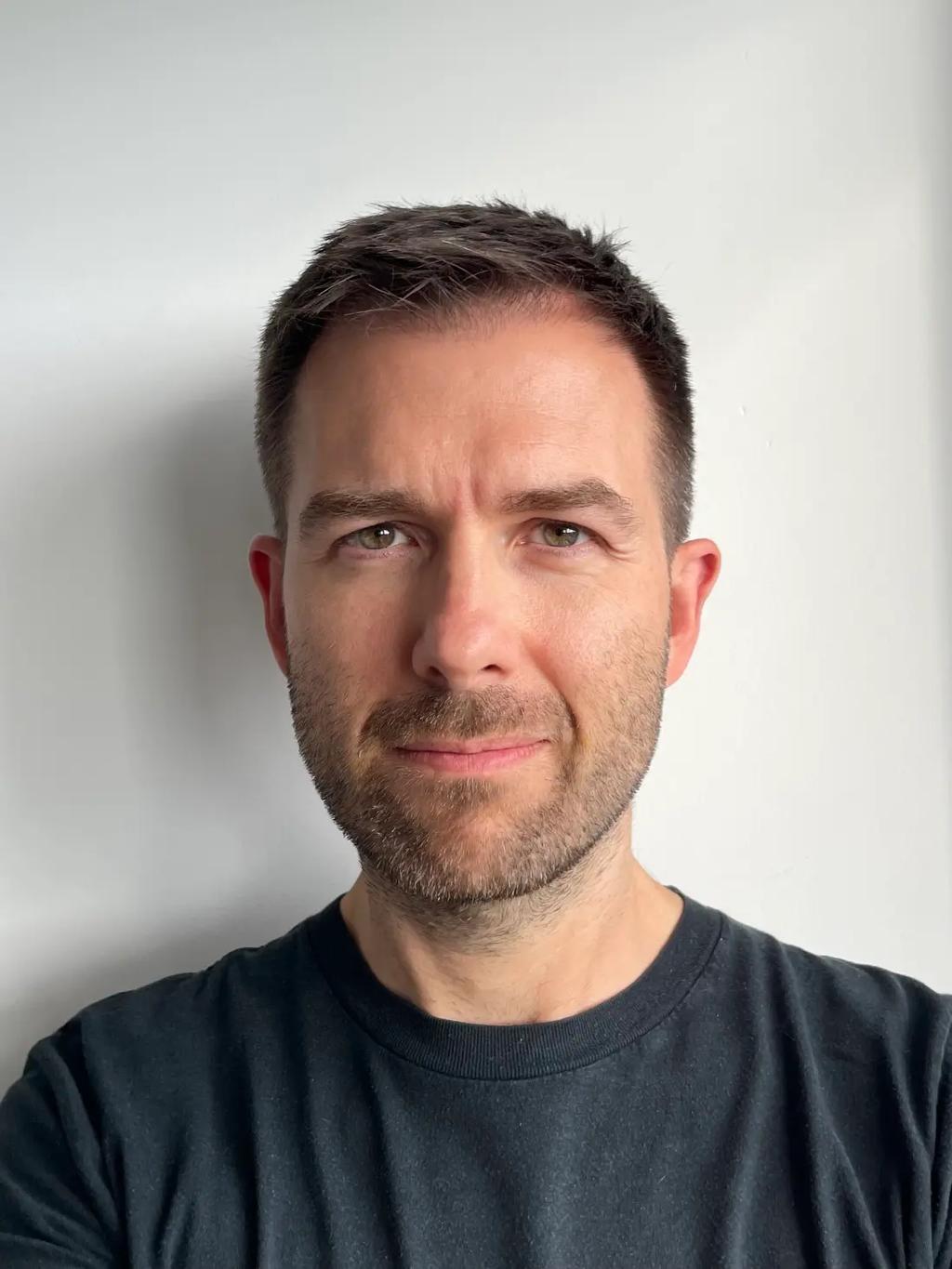 Man with short brown hair, light beard, and a black t-shirt standing against a plain white wall.