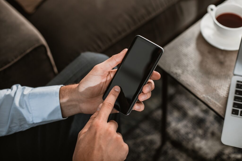 mobile app addiction - pair of hands using a smartphone, the smartphones screen is blank and in the background there is a sofa, coffee cup and a laptop