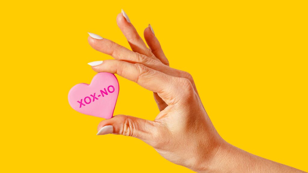A hand with white manicured nails holds a small pink heart-shaped object with "XOX-NO" written on it in a darker pink colour". The image is set against a bold yellow background. 