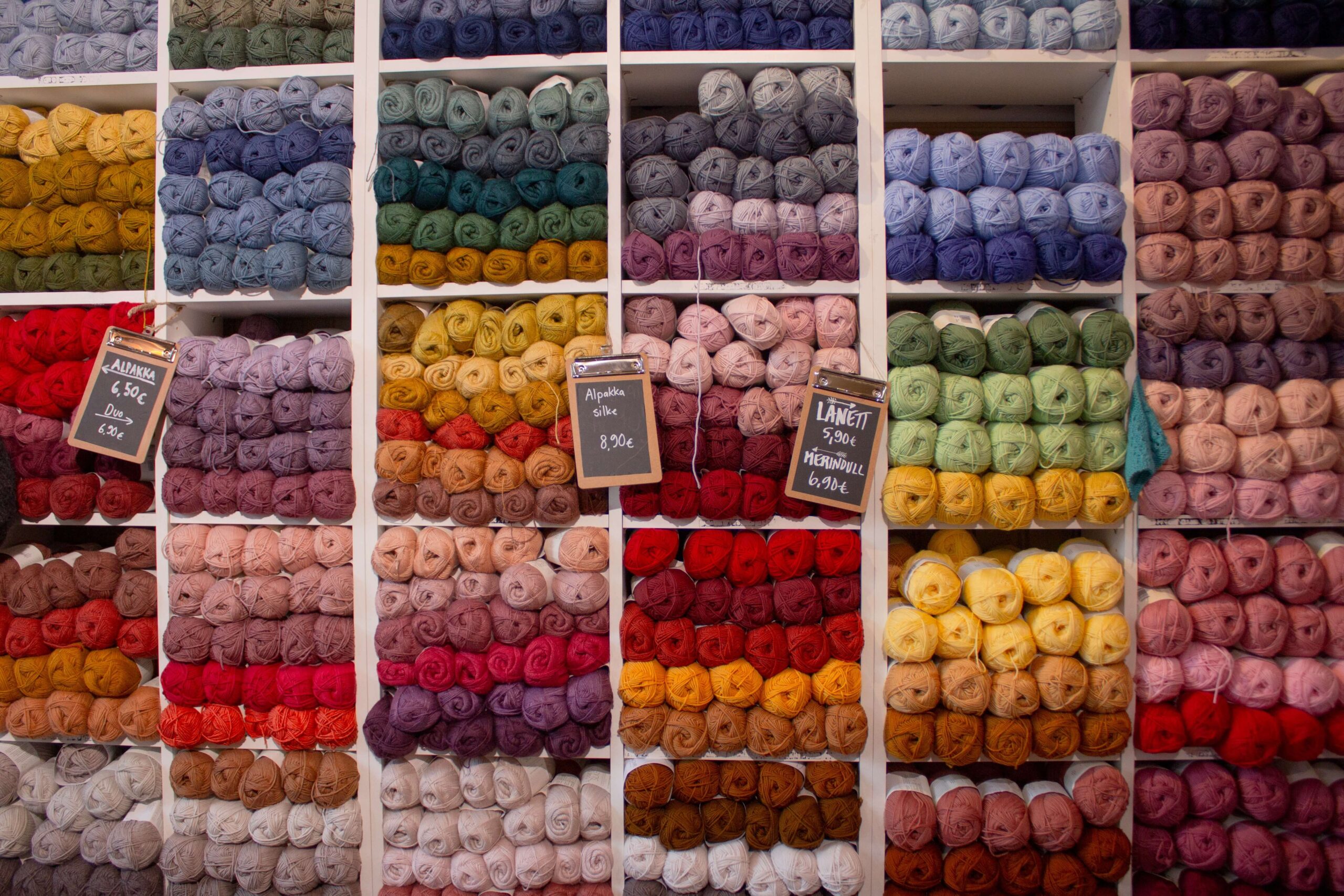 Shelves of balls of wool on display in a shop. Hundreds of balls of wool arranged into groups of different bright and pastel colours. Three small blackboards hand from the shelves displaying the prices.