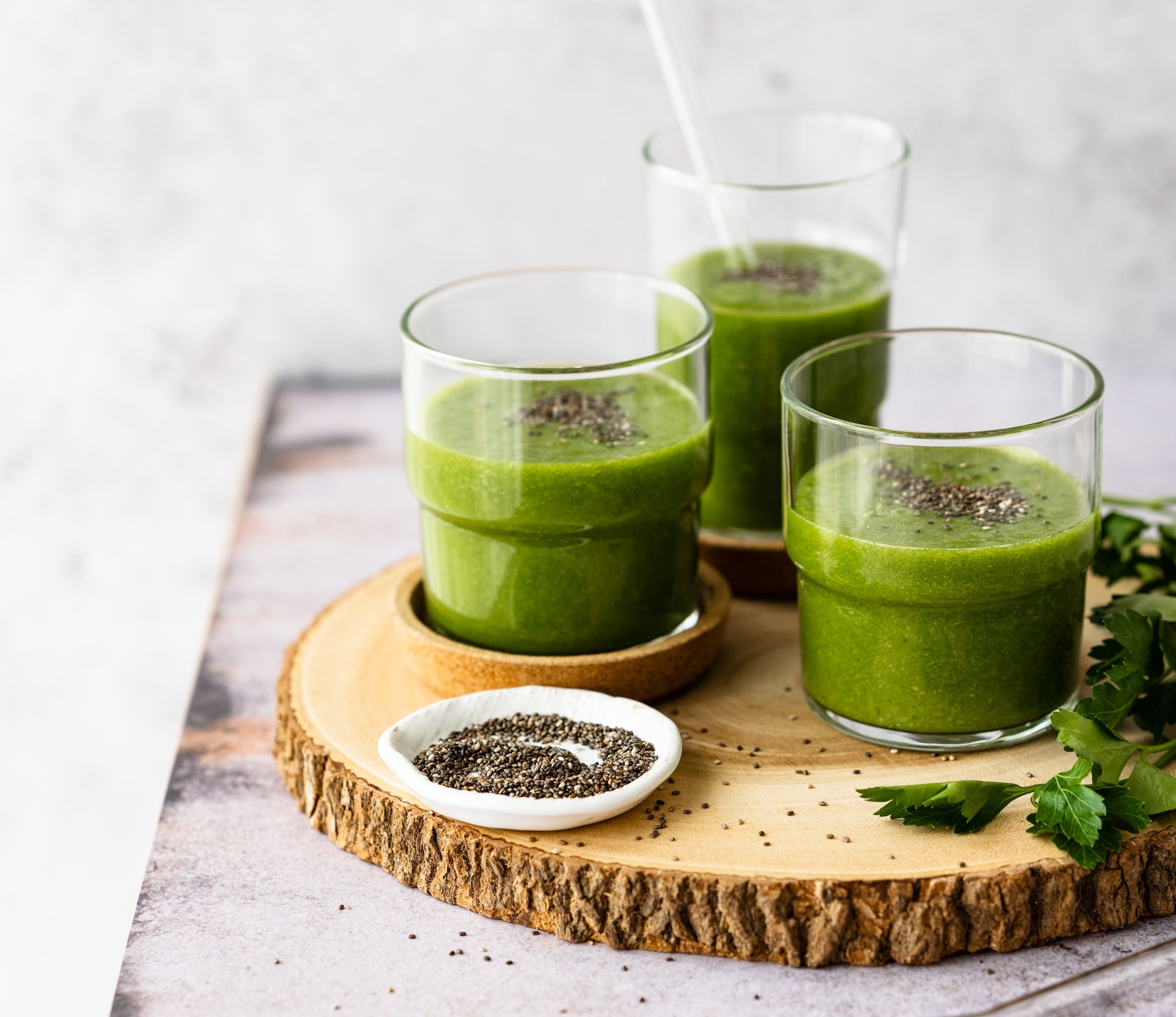 Three glasses of green fruit and vegetable smoothies placed on a wooden board. Chia seeds have been sprinkled into the smoothies and across the board. The board is dressed with some parsley leaves.