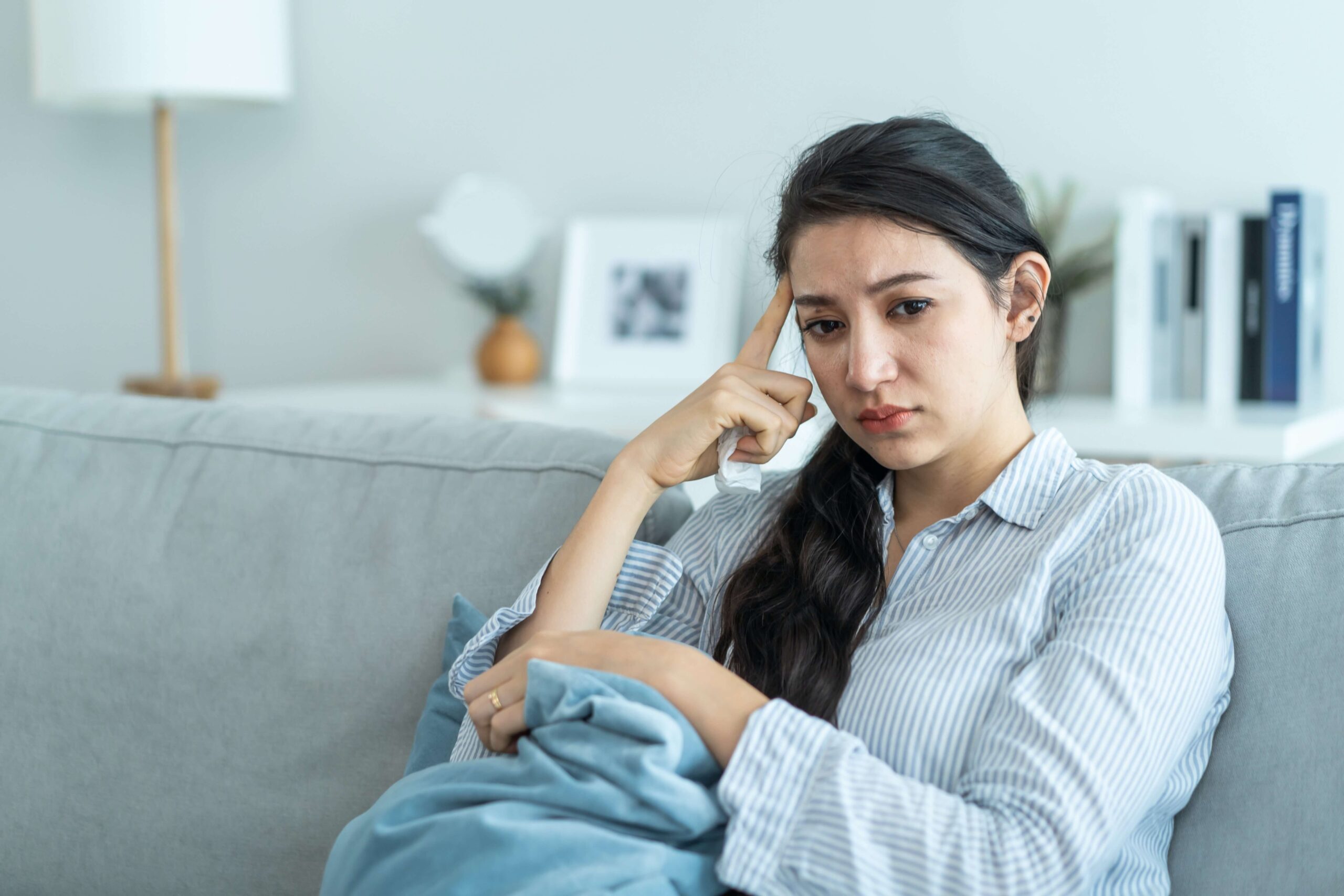 A person with the characteristics of a woman is sitting on a light grey sofa looking troubled, clutching a cushion to one side. Their gaze is down towards the floor. Posed by model.