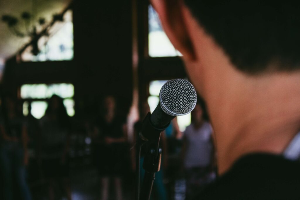 Person with back to the camera facing a microphone and beyond the microphone a small crowd are looking towards the person.