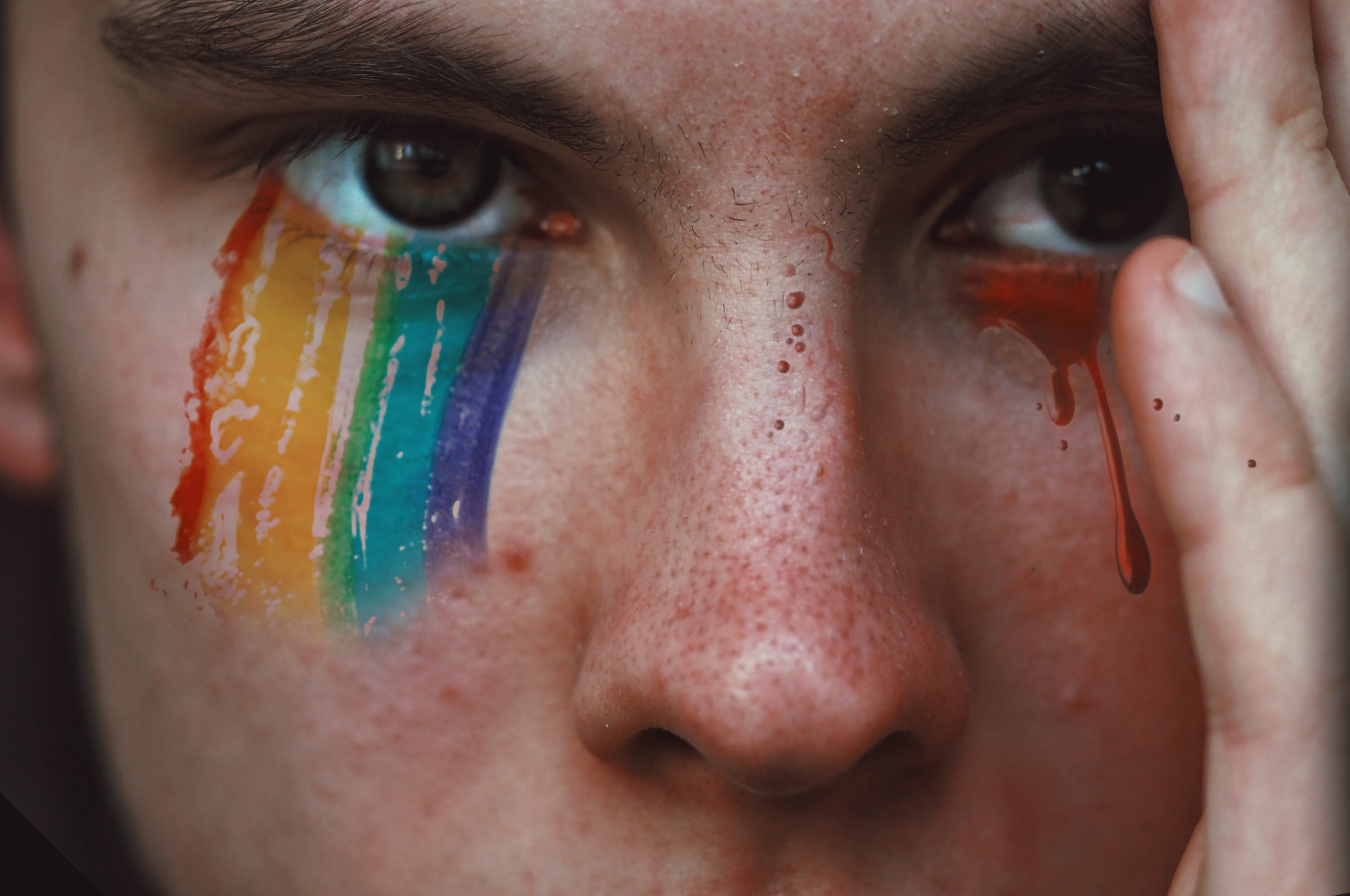 Close up of the face of a person, we see just their eyes and nose, with their hand shielding part of their face. A graphical overlay places rainbow-coloured mascara under one eye and blood droplets under the other. Posed by model.