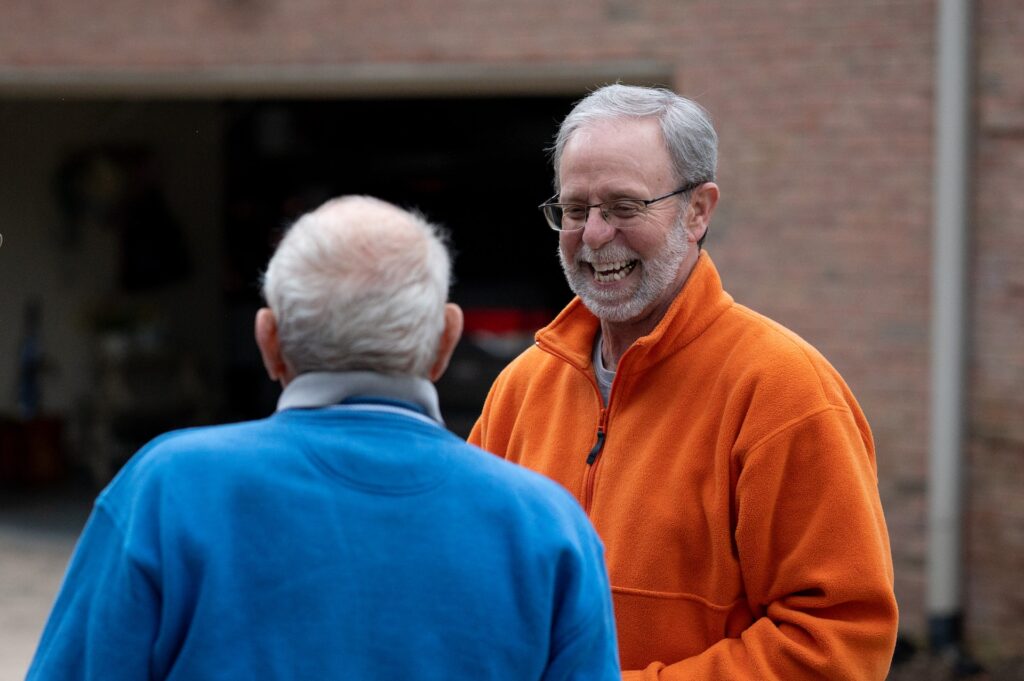 Two men with grey hair facing one another. One in a blue jacket with his back to us. The other in an orange jacket is smiling towards him