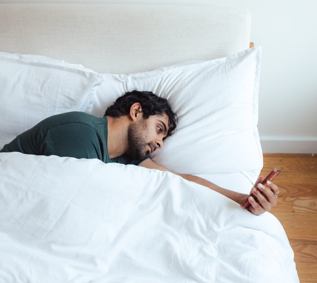 A person checking their smartphone in bed. They are lying on their side, wearing a dark t-shirt, in a white bed. Digital addiction and sleep disturbance.