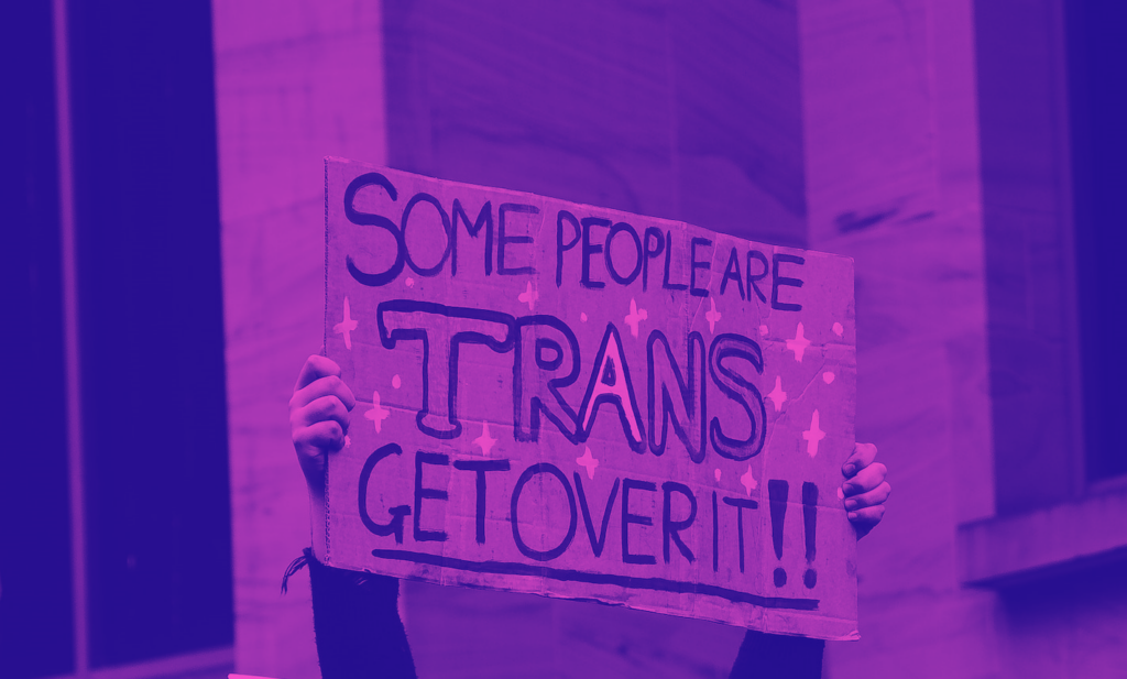 Sign being held above someone's head saying "some people are trans get over it!!"