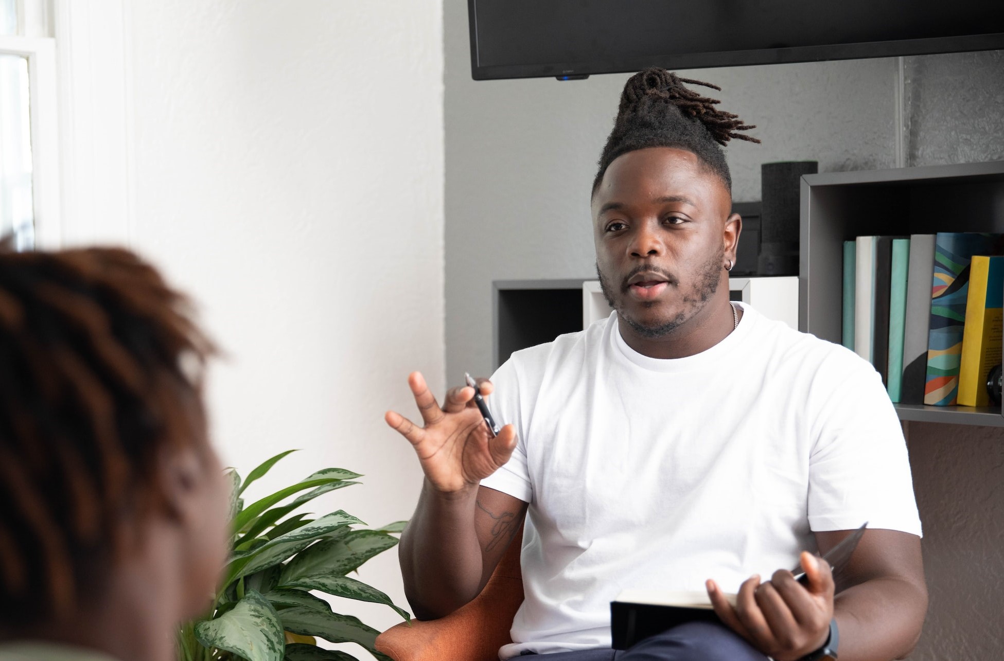 A man in a white t-shirt sits in an office, holding a notebook in one and a pen in the other. He is looking towards another person, we can see just the back of their head. It appears that they are in some sort of therapy session.