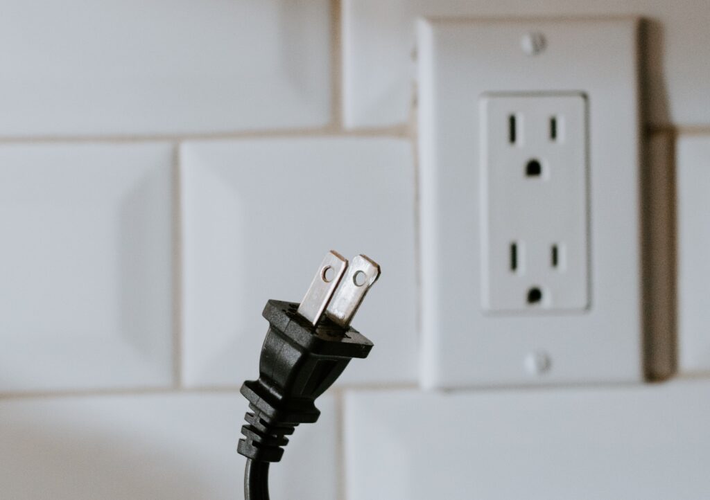 A black electrical plug, probably from an appliance in the USA, is positioned in front of a North American electrical wall plug socket. 
