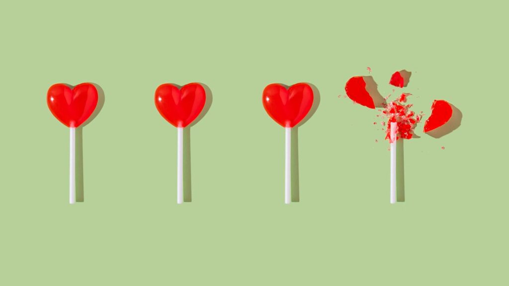 Picture of four heart-shaped red lollipops (hard sugar candy) on white sticks in a row, against a green background. The 4th lollipop is broken into little pieces.