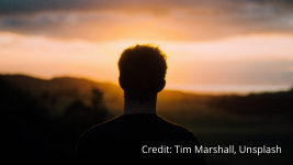gay-self-acceptance Silhouette of a person with short hair standing outdoors, facing a sunset over a landscape, with the sky glowing orange and clouds above. Credit: Tim Marshall, Unsplash.