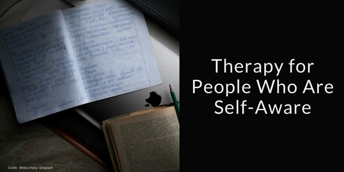 Open notebook with handwritten text in blue ink, a pencil, and an open book placed on a laptop, all on a desk. The left side of the image shows these items in soft light, while the right side features a black background with the white text: "Therapy for People Who Are Self-Aware." The photo credit in small text reads: "Credit: Misha Vrana, Unsplash